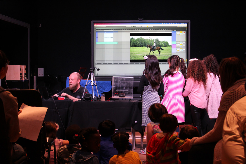 A man presenting on a screen, using a computer to record sounds being made by a group of young girls wearing pink and blue dresses, standing to his left, in front of an audience with families.